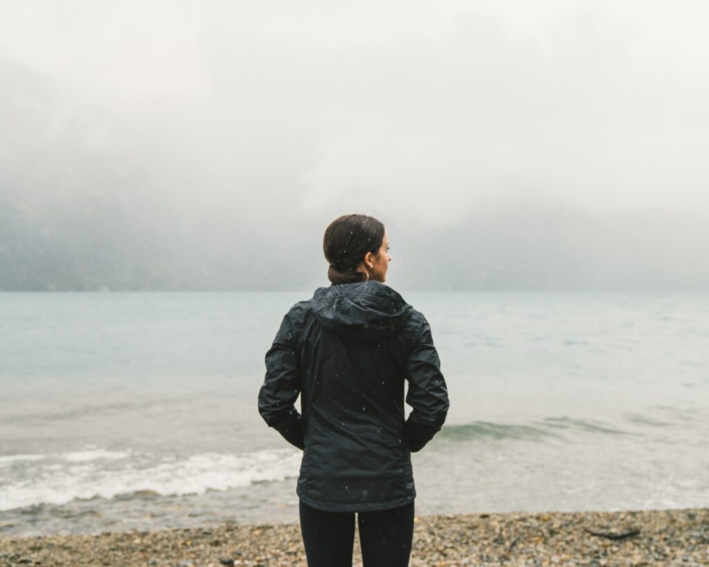 A brunette woman stands on a rainy beach looking out at the water during off season.