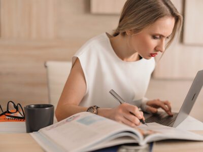 A blonde woman sits at a desk with a notebook and computer while researching branding for her short-term rental.