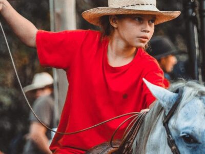 Child in a red t-shirt and cowboy hat riding a horse while holding a lasso at a rodeo.