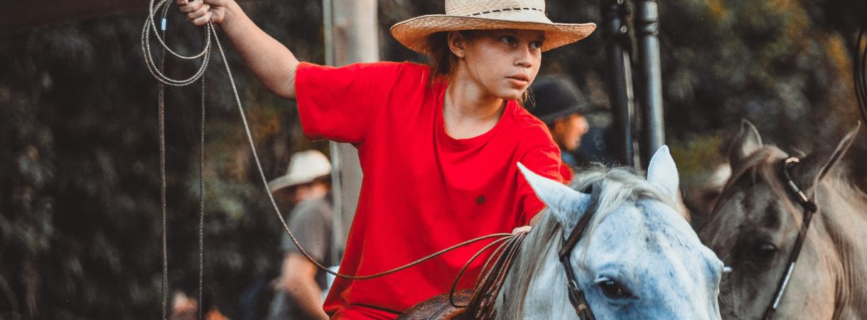 Child in a red t-shirt and cowboy hat riding a horse while holding a lasso at a rodeo.